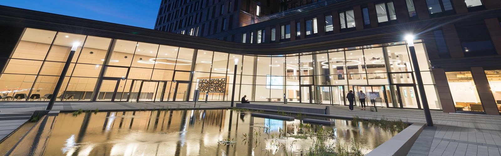 University building at night. Thr windows are huge and displaying the light from the inside. A large fountain is in the courtyard of the building.